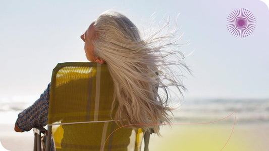 mulher mais velha sentada em uma cadeira de frente para o mar com seus cabelos longos esvoaçantes