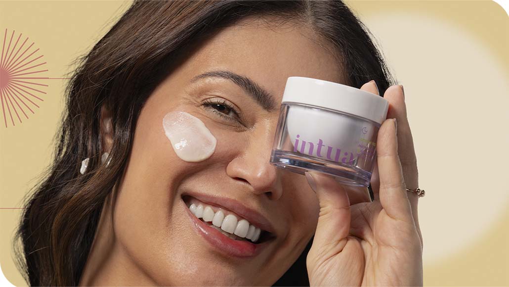 mulher segurando um pote de Creme Nutritivo Facial da Intua com um pouco do produto aplicado em sua bochecha