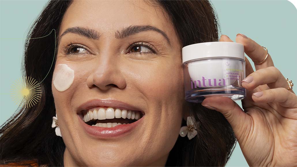 mulher segurando um pote do Creme Nutritivo Facial da Intua com um pouco do produto aplicado em seu rosto