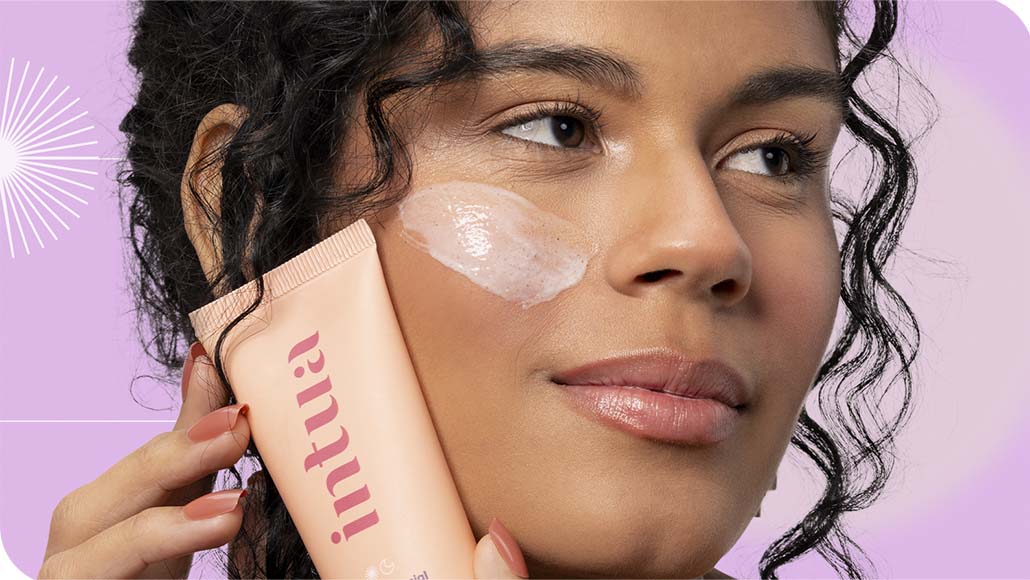 mulher segurando o Esfoliante Facial da Intua contra o rosto, com um pouco do produto aplicado em sua bochecha