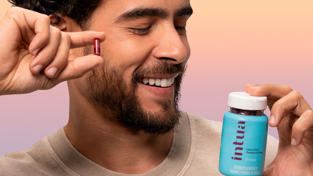 Homem segurando uma cápsula do Nutricosmético Cabelos & Unhas da Intua Beauty perto do rosto e com a outra mão segura um pote do produto.
