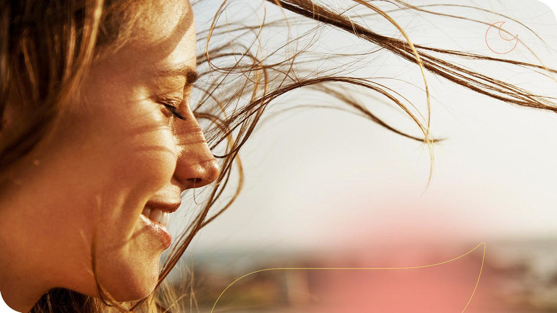 mulher sorrindo com os olhos fechados com seus cabelos compridos esvoaçantes em um campo aberto sob o sol